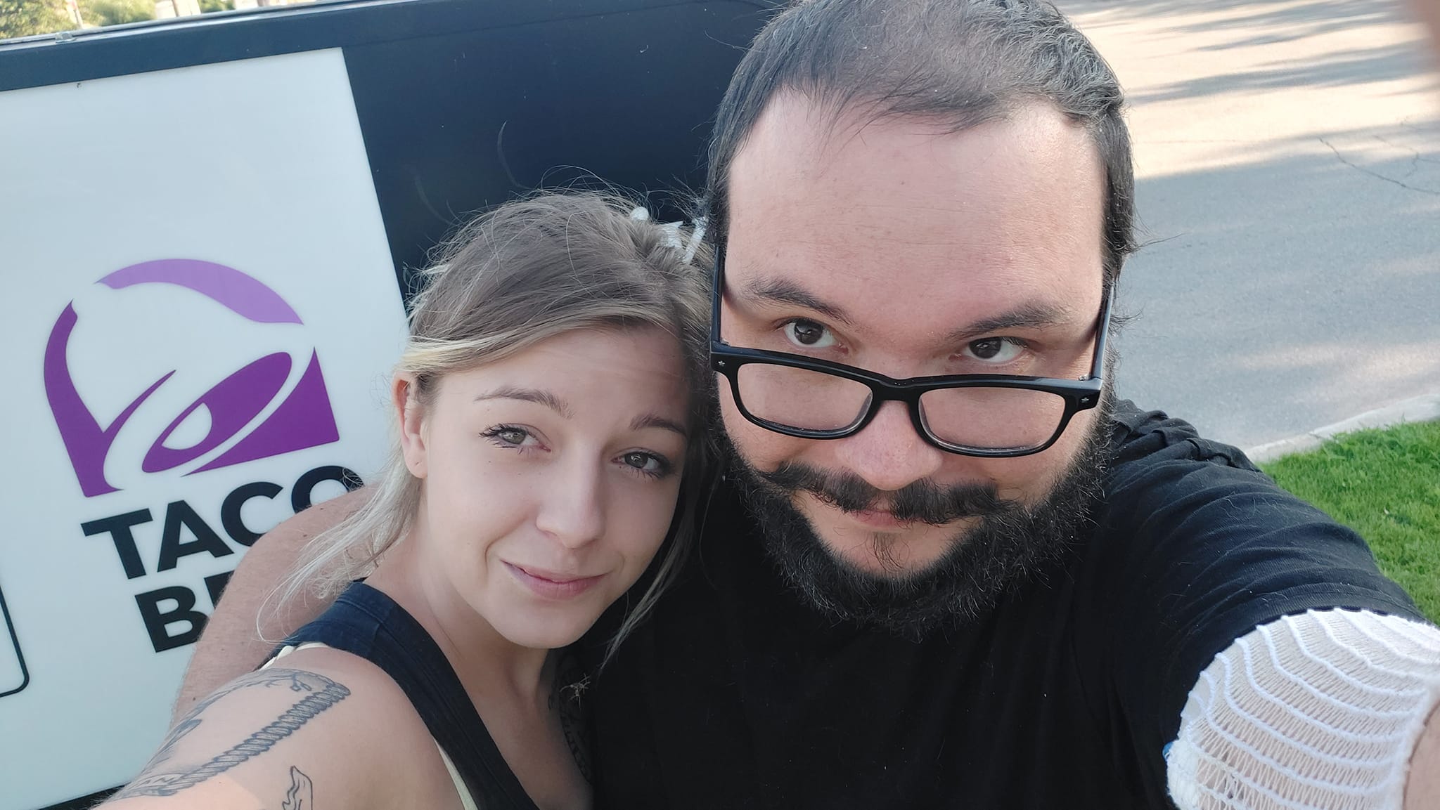 A man short hair and bushy facial hair hugs a small blond woman next to the KFC/Taco Bell sign